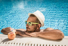 Portrait Of A Handsome Man Resting At The Edge Of A Swimming Pool. Model Is Wearing Hat And Sunglasses. Tropical Drink With Drinking Straw And Paper Umbrella Next To Him.  Summer Vacation Concept. 