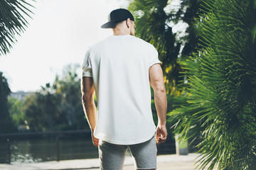 Wall Mural - Photo Bearded Muscular Man Wearing White Blank t-shirt, snapback cap and shorts in summer time. Green City Garden Park Background. Back view. Horizontal Mockup