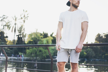 Poster - Image Bearded Muscular Man Wearing White Blank t-shirt, snapback cap and shorts in summer vacation. Relaxing time near the lake. Green City Garden Park Sunset Background.Back view.Horizontal Mockup