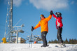 © anatoliy_gleb - Man and woman holding hands, smiling looking at each other standing with skis on mountain top and giving each other a high five at a winter resort with ski lifts and blue sky in background