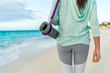 © Maridav - Fitness woman carrying yoga mat with strap on beach going to class training. Closeup of sports equipment, back view of fit athlete in activewear showing fashion leggings and turquoise hoodie.
