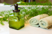 Pump Green Glass Bottle With Liquid Castile Soap. Rolled Green Towels In A Spa Setting. Green Plant Decor In Background. Bathroom White Countertop.