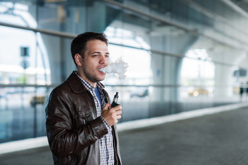 Man with a bristle smoking e-cigarette vaporizer box mode outdoors near the airport terminal before flight