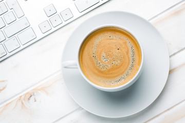 Coffee in a white cup and saucer standing on a wooden table next to a keyboard