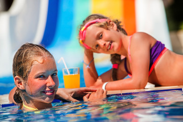 Wall Mural - Two children on water slide at aquapark lie on edge of swimming pool and drinking cold orange drink.  Children lie on edge of swimming pool. Summer holiday.Outdoor.