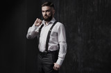 Fototapeta  - Attractive bearded man in a white shirt and suspenders standing near dark wall. He is strong, courageous and serious. He holds the collar of his shirt. Dark room, night and harsh shadows.