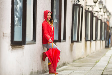 Wall Mural - Portrait of a beautiful young woman in a red hooded raincoat leaning on the wall and looking at camera.