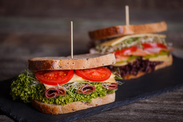 Two Sandwiches with cereals bread with lettuce, slices of fresh tomatoes and sweet pepper, ham, prosciutto, salami, cheese  and  onion sprouts on dark marble background