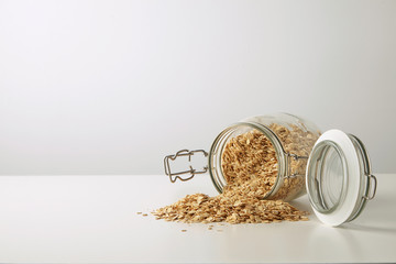 Full opened rustic jar with healthy rolled oats spread out isolated in center on white table side view Breakfast muesli set