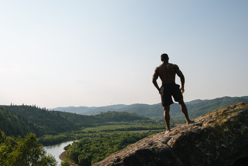 Silhouette of african american athletic man with naked torso posing on the rock. Green mountain nature background