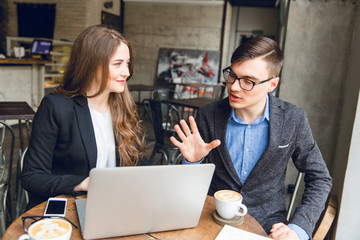 Two colleagues discuss something near laptop. Man in blue shirt and grey jacket describes something with hand gestures. Woman with long hair wearing black jacket and white blouse listens and smiles.