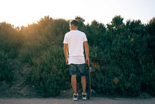 A Man Wearing A Blank White T-shirt And Shorts Is Standing With His Back To The Camera. A Skater Boy With A Skateboard Is Looking Aside On A Bushy Background.