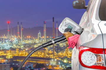 Bronze car at gas station being filled with fuel and the blurred oil refinery with tube and oil tank along night sky at Si Racha District background, Transportation Concept, selective focus