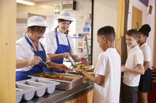 Two Women Serving Food To A Boy In A School Cafeteria Queue