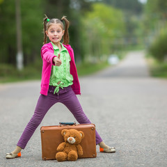 Little girl in bright clothes standing on the road with a suitcase and a Teddy bear.