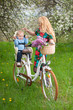 © anatoliy_gleb - Beautiful blonde woman with long hair and red lips in flowered dress holding baby in bicycle chair, against the background of blooming trees, dandelions and fresh greenery in spring garden