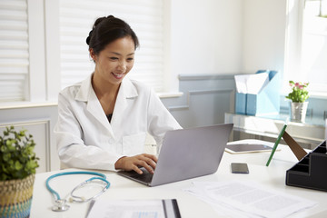 Female Doctor Sitting At Desk Working At Laptop In Office