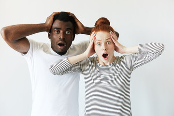 Wall Mural - Surprised African male and redhead Caucasian female wearing casual clothes looking at the camera in shock, surprised with sale prices, holding hands on head, mouth wide open. Mixed race relationships