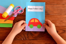 Child Holds A Greeting Card In His Hands. Kids Crafts For Father's Day. Card With Message Happy Father's Day. Holiday Gift For Dad. Sheets Of Colored Paper, Scissors, Glue Stick. Wooden Background
