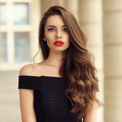 Wall Mural - Close up portrait of young beautiful woman with long brunette curly hair posing against architectural background and looking at you