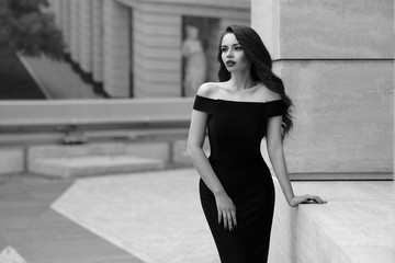 Black and white portrait of young beautiful elegant woman in black dress. Pretty sensual girl with long curly hair posing outdoors at city street