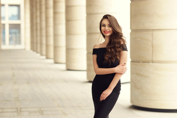 Young elegant girl posing at city street. Pretty beautiful business woman in elegant black dress against city background.