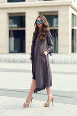 Outdoor dynamic fashion portrait of young beautiful stylish woman in black dress, grey coat and sunglasses walking on a windy day against city background