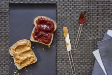 Preparing Peanut Butter And Strawberry Jam Sandwich On Black Flat Plate With Dirty Spoon And Knife Aside And Folded Blue And Gray Napkin On Wrap Textured Background