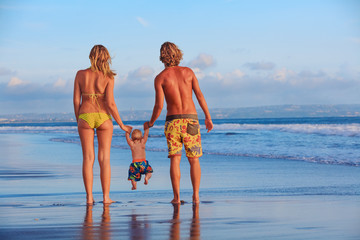 Wall Mural - Happy family - father, mother, baby son hold hands, swim with fun, walk along sunset sea surf on black sand beach. Travel, active parents lifestyle, people activity on summer vacations with children.
