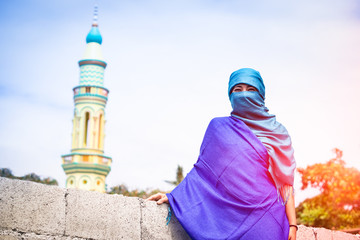 Wall Mural - Young muslim woman standing with traditional islamic clothing - Portrait of asian girl with veiled face on minarets background - Concept of world religions