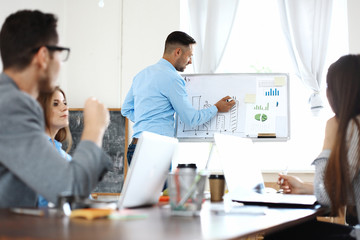 Wall Mural - Handsome young man standing near whiteboard and pointing on the chart while his coworkers listening and sitting at the table