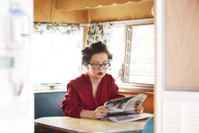 Woman In Bathrobe And Curlers Reading Newspaper In Camper Van