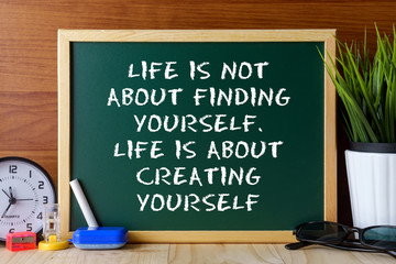 Word quote LIFE IS NOT ABOUT FINDING YOURSELF,LIFE IS ABOUT CREATING YOURSELF written on green chalk board on wooden table.