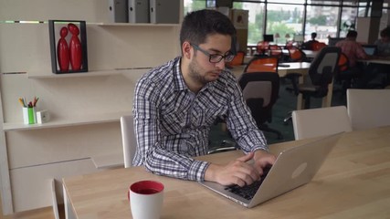 Fototapete - Handsome man mixed race typing on the computer at his working place. On the background colleagues work with laptop. Brunet with eyeglasses on his face. Behind young male wooden sh