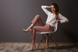 © Africa Studio - Beautiful young woman in shirt and pink pants sitting on chair on dark grey wall background