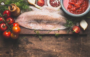 Raw fish fillet with frish herbs, tomatoes and sauce on dark rustic cutting board on dark aged wooden background, top view, border. Pollack fish cooking