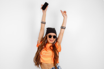 beautiful woman in sunglasses wearing in black hat and orange T-shirt listening music and dancing near white wall, holding a cell phone in hand, hands raised up