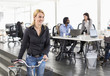 © Maskot - Mature businesswoman looking away while holding bicycle with colleagues working in background at office