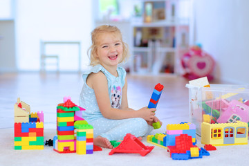 Little preschooler girl building house from plastic blocks. Lovely laughing child, blonde girl of preschool age playing with colorful bricks sitting on carpet in a sunny room at home or kindergarten