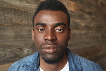 Close up portrait of dark-skinned man looking seriously at camera on wooden background. Thoughtful and peaceful facial expression, relaxed and regular mimics of African person with little beard.