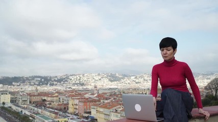Poster - Female typing on the laptop looking on the city view. Nice town Cote d'Azur. Professional businesswoman at holiday vacation. Attractive girl with short hair wearing in casual red pullover and blue