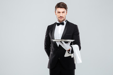 Handsome yong waiter in tuxedo and gloves holding empty tray