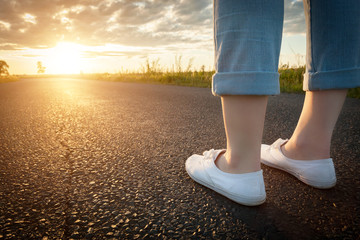 woman in white sneakers standing on asphalt road towards sun. travel, freedom concepts.