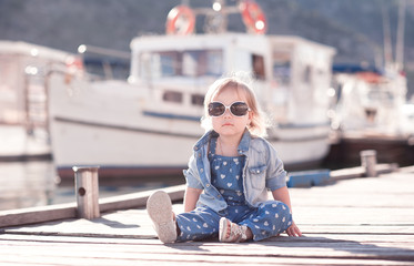 Stylish kid girl wearing trendy denim clothes sitting on wooden sea pier outdoors. Looking at camera. Childhood.