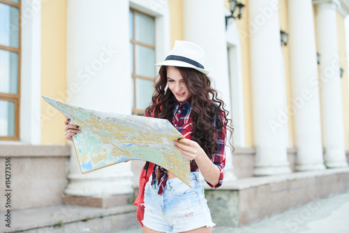 Fotografía Tourist girl in a hat holding a map and looking at her against the building with