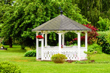 Lovely White Gazebo With Red Flowers Hanging From A Basket. Trees And Shrubs In The Background. Fine Garden With Green Grass.