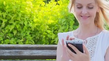 High Quality 10bit Footage Of Happy Smiling Girl Using A Smart Phone In A City Park. Made From 14bit RAW.