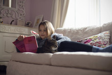 Young Girl Lying On Sofa At Home Reading Book With Pet Cat