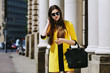 Outdoor portrait of young beautiful lady walking on the street. Model wearing sunglasses & stylish yellow summer dress. Girl looking down. Female fashion concept. City lifestyle. Sunny day. Waist up
