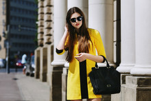 Outdoor Portrait Of Young Beautiful Lady Walking On The Street. Model Wearing Sunglasses & Stylish Yellow Summer Dress. Girl Looking Down. Female Fashion Concept. City Lifestyle. Sunny Day. Waist Up